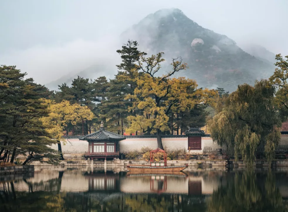 Koreansk hus ved vannet. Et fjell stikker opp bak trærne i bakgrunnen.